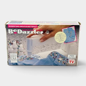 May include: A BeDazzler rhinestone and stud setter kit in a rectangular box. The packaging features a white handheld tool applying colorful rhinestones to blue denim fabric. Text on the box includes BeDazzler, Made in U.S.A., and U.S. Patents 3960307, D239320.