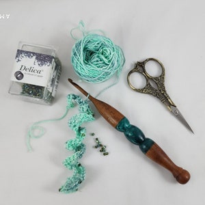 May include: A close-up of a crochet project with green and blue yarn, a wooden crochet hook, a small container of seed beads, and a pair of gold-colored scissors.
