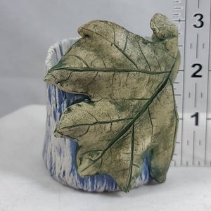 May include: A small ceramic planter with a blue and white glaze. A green and brown leaf is attached to the side of the planter.