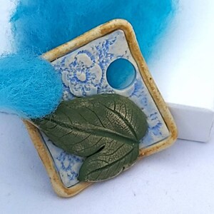 May include: Square ceramic tile with a blue and white floral design, a green leaf, and a blue circle in the center. The tile is surrounded by a beige border.