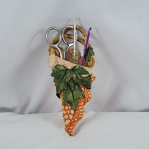 May include: A ceramic wall-mounted holder shaped like a grapevine with green leaves. It has a silver pair of scissors and a purple crochet hook inside.