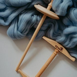 May include: Two wooden drop spindles with a light blue wool roving background. One spindle is upright, the other is angled. The spindles have a natural wood finish. The word "Kromski" is visible on one spindle.