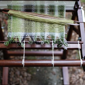 May include: A wooden loom with a green, white, and black woven fabric on it. The loom is set up with white string and a wooden dowel.