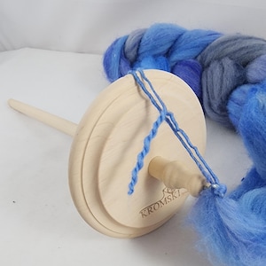May include: A wooden Kromski spinning drop spindle with a light blue yarn attached. A braid of blue and gray wool fiber is next to the spindle. The spindle has a wooden shaft and a round wooden whorl.