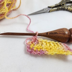 May include: A wooden crochet hook, labeled "4 mm", is shown with a partially completed crochet project. The yarn is a blend of yellow, pink, and white. Vintage scissors are in the background. The project is in progress.