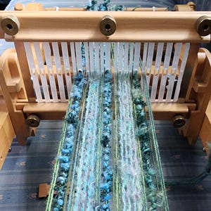 May include: A close-up of a loom with a woven fabric in shades of blue, green, and white. The fabric is made with a variety of yarns, including some with a fuzzy texture.