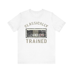 Könnte beinhalten: Weißes T-Shirt mit einem verblichenen Grafikdesign eines klassischen Videospiel-Controllers. Der Text "CLASSICALLY TRAINED" ist über und unter dem Controller-Bild in einer Vintage-Schriftart aufgedruckt. Ein lässiges T-Shirt für Gamer.