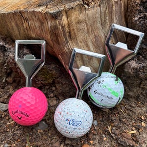 Peut inclure: Trois décapsuleurs dotés de têtes en métal argenté fixées sur des balles de golf. L'une est une balle Callaway rose unie, une autre est une balle Vice blanche mouchetée, et la troisième est une balle TaylorMade blanche avec des éclaboussures.