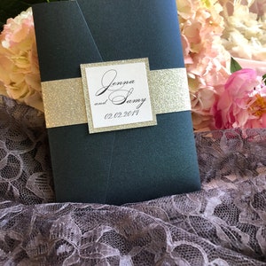 May include: A dark blue wedding invitation with a white square overlay. The square has the text "Jenna and Sammy 02.02.2019" in black cursive font. The invitation is decorated with a gold glitter ribbon.