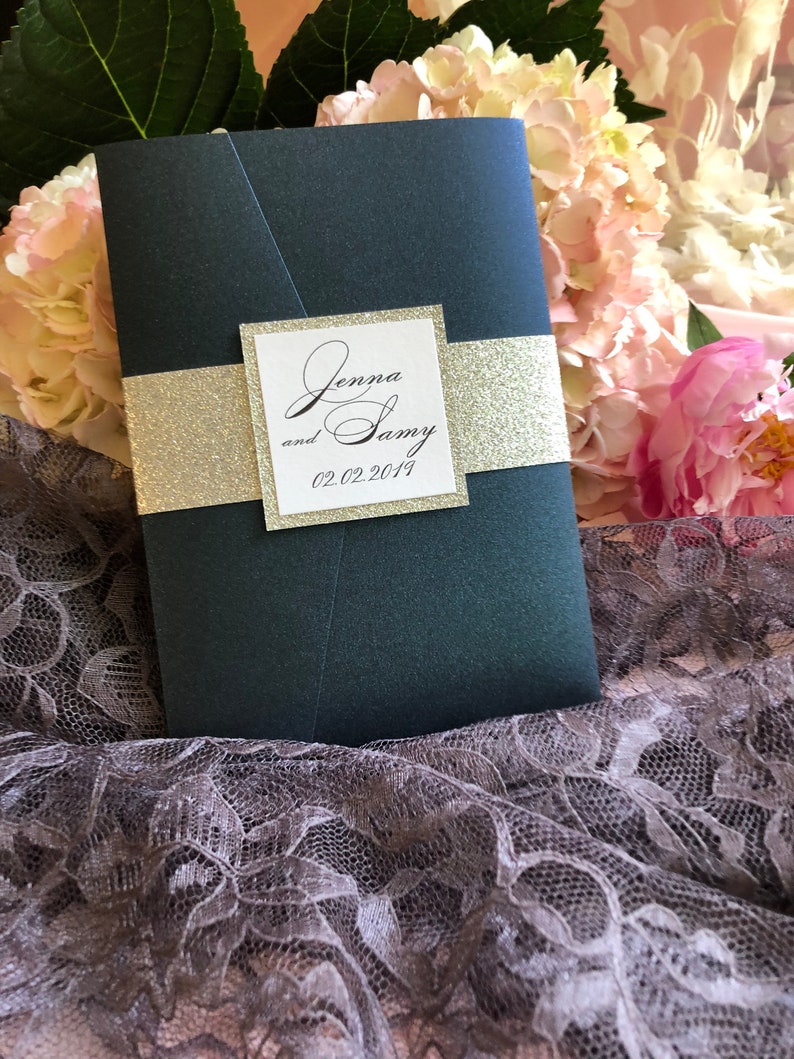 May include: A dark blue wedding invitation with a white square overlay. The square has the text "Jenna and Sammy 02.02.2019" in black cursive font. The invitation is decorated with a gold glitter ribbon.