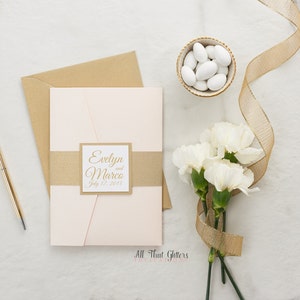 May include: A blush pink wedding invitation with a gold envelope and a gold ribbon. The invitation has a square label with the text "Evelyn and Marco July 17, 2015" in gold script.