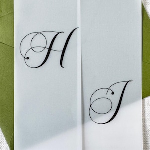 Puede incluir: Primer plano de una invitaci&oacute;n de boda con las letras "H" y "J" en elegante escritura negra. La invitaci&oacute;n est&aacute; sobre papel transl&uacute;cido, con un sobre verde en el fondo. El dise&ntilde;o es simple y sofisticado.