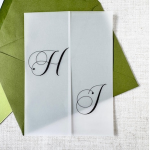 Puede incluir: Una tarjeta de invitaci&oacute;n transl&uacute;cida plegada, con las letras "H" y "J" en elegante escritura negra. La tarjeta est&aacute; colocada frente a sobres verdes, sugiriendo un tema de boda o evento especial.