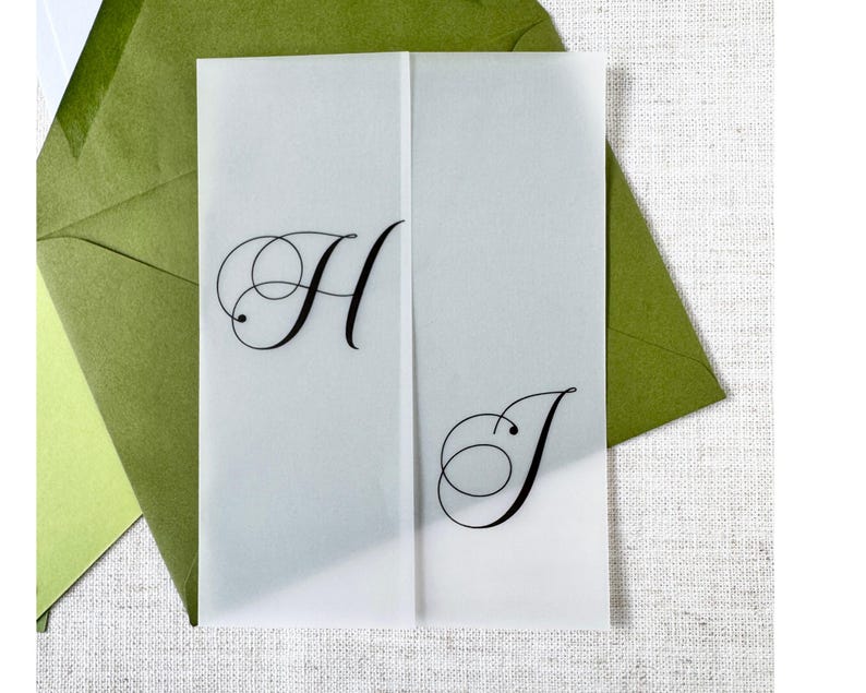 Puede incluir: Una tarjeta de invitaci&oacute;n transl&uacute;cida plegada, con las letras "H" y "J" en elegante escritura negra. La tarjeta est&aacute; colocada frente a sobres verdes, sugiriendo un tema de boda o evento especial.