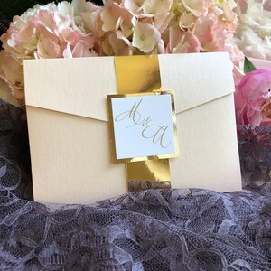 May include: A white wedding invitation with a gold envelope and a gold ribbon. The invitation has the initials "M & A" written in gold calligraphy.