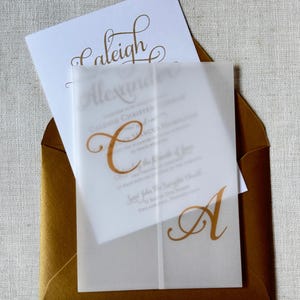 May include: A gold-colored envelope holds a white invitation card with gold calligraphy. A translucent overlay features the letter "C" and the letter "A" in gold. The invitation is for a wedding, with the names "Caleigh" and "Alexander" visible.