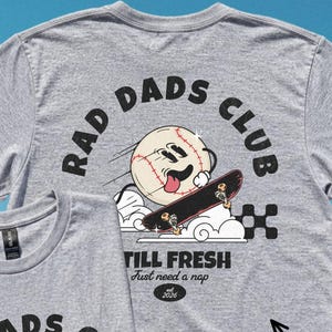 Op de afbeelding: Een sportgrijs t-shirt met een afbeelding van een cartoon honkbal op een skateboard met de tekst RAD DADS CLUB, STILL FRESH, Just need a nap en est. 2026. Het ontwerp bevat een dambordpatroon en wolken. Een tekstvak vermeldt FRONT OR BACK PRINT SELECT INSIDE!
