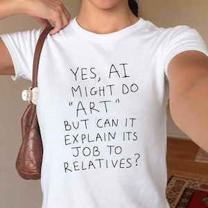 May include: A white short-sleeved t-shirt featuring black hand-drawn style text that reads: YES, AI MIGHT DO "ART" BUT CAN IT EXPLAIN ITS JOB TO RELATIVES? The shirt has a crew neckline and a fitted silhouette, worn with a brown shoulder bag.