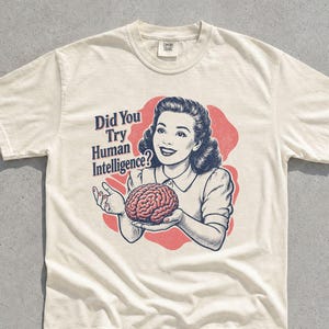 Op de afbeelding: Een ivoorkleurig t-shirt met korte mouwen en een grafische illustratie van een persoon die een brein vasthoudt. De tekst luidt, Did You Try Human Intelligence? Het ontwerp gebruikt een rode achtergrondvorm achter de figuur met donkerblauwe inkt.