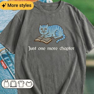 K&ouml;nnte beinhalten: Ein anthrazitfarbenes Kurzarm-T-Shirt mit einer Grafik einer blau gestreiften Katze, die mit einem offenen Buch daliegt. Unter der Katze steht der Text, Just one more chapter. Das T-Shirt hat einen Rundhalsausschnitt und eine Vintage-Stoffoptik.