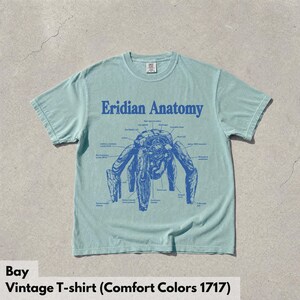 May include: A light green short-sleeve t-shirt featuring a blue graphic print of a creature labeled Eridian Anatomy. The design includes various anatomical labels with lines pointing to specific parts of the creature, centered on the chest.