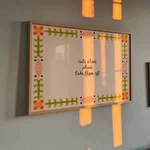 May include: A rectangular wall sign in a light wood frame featuring a white background with a decorative border of green leafy vines, orange geometric shapes, and pink flowers. The text in the centre reads, "cute shoes please take them off."