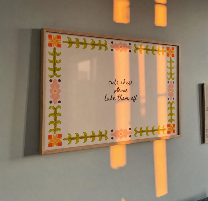 May include: A rectangular wall sign in a light wood frame featuring a white background with a decorative border of green leafy vines, orange geometric shapes, and pink flowers. The text in the centre reads, "cute shoes please take them off."