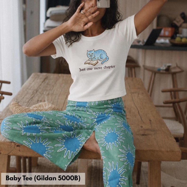 K&ouml;nnte beinhalten: Ein wei&szlig;es kurz&auml;rmeliges Baby-T-Shirt mit einer Grafik einer blau gestreiften Katze, die auf einem offenen Buch liegt, und dem darunter gedruckten Text Just one more chapter. Dazu wird eine gr&uuml;n-blaue Hose mit Sonnenstrahlenmuster getragen.