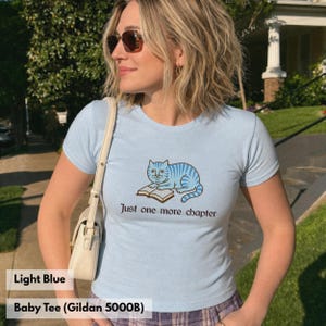 K&ouml;nnte beinhalten: Ein hellblaues T-Shirt mit kurzen &Auml;rmeln und einer Grafik einer blau gestreiften Katze, die auf einem offenen Buch liegt. Unter der Katze steht der Text Just one more chapter in dunkler Schrift. Das T-Shirt hat einen Rundhalsausschnitt.