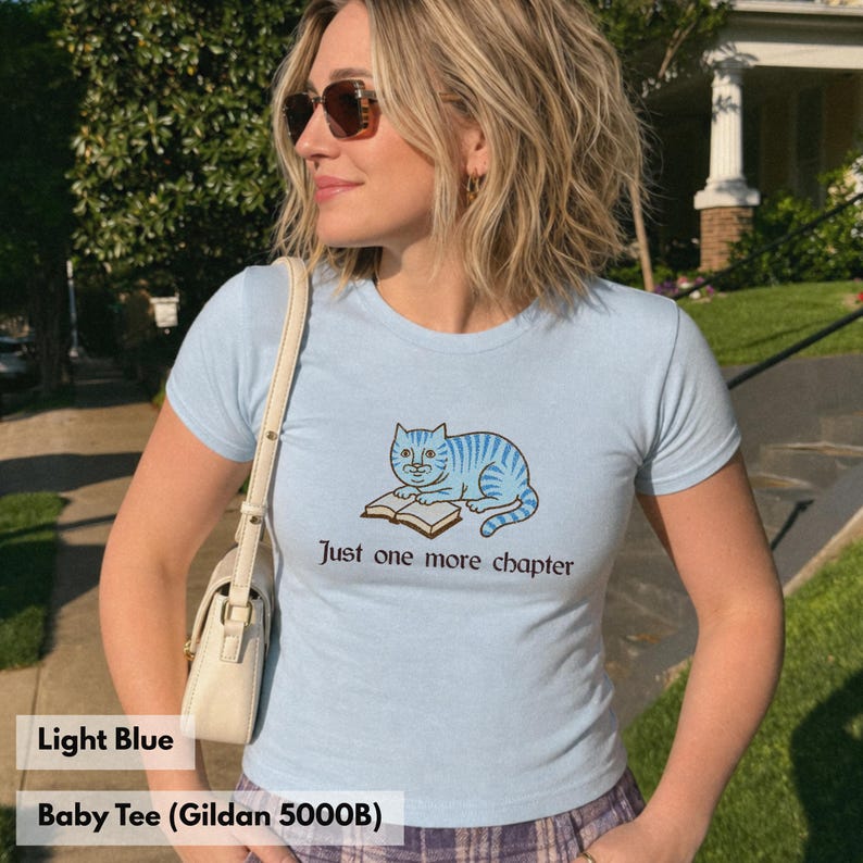 K&ouml;nnte beinhalten: Ein hellblaues T-Shirt mit kurzen &Auml;rmeln und einer Grafik einer blau gestreiften Katze, die auf einem offenen Buch liegt. Unter der Katze steht der Text Just one more chapter in dunkler Schrift. Das T-Shirt hat einen Rundhalsausschnitt.