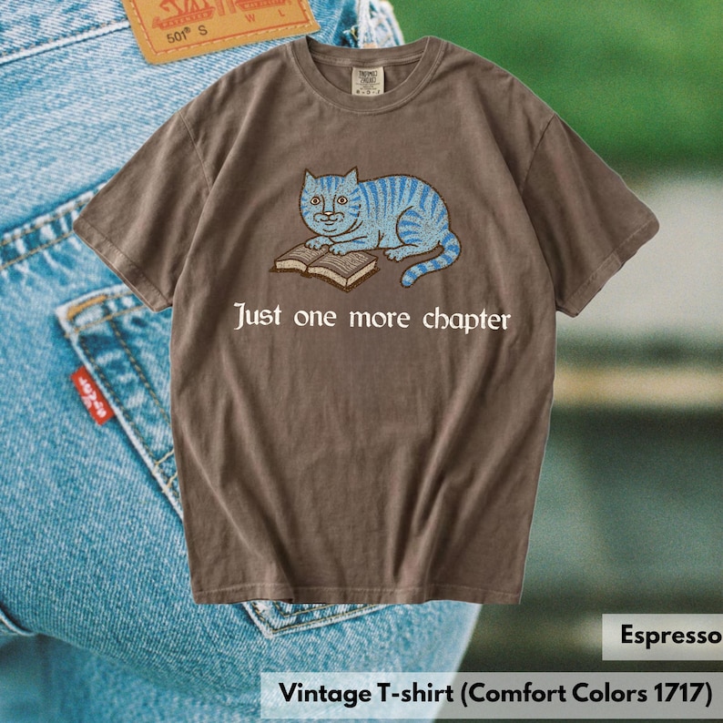 K&ouml;nnte beinhalten: Ein espresso-braunes Kurzarm-T-Shirt mit einer Grafik einer blau gestreiften Katze, die auf einem offenen Buch liegt. Unter der Katze steht der Text Just one more chapter in einer wei&szlig;en Serifenschrift. Das T-Shirt hat einen Rundhalsausschnitt.