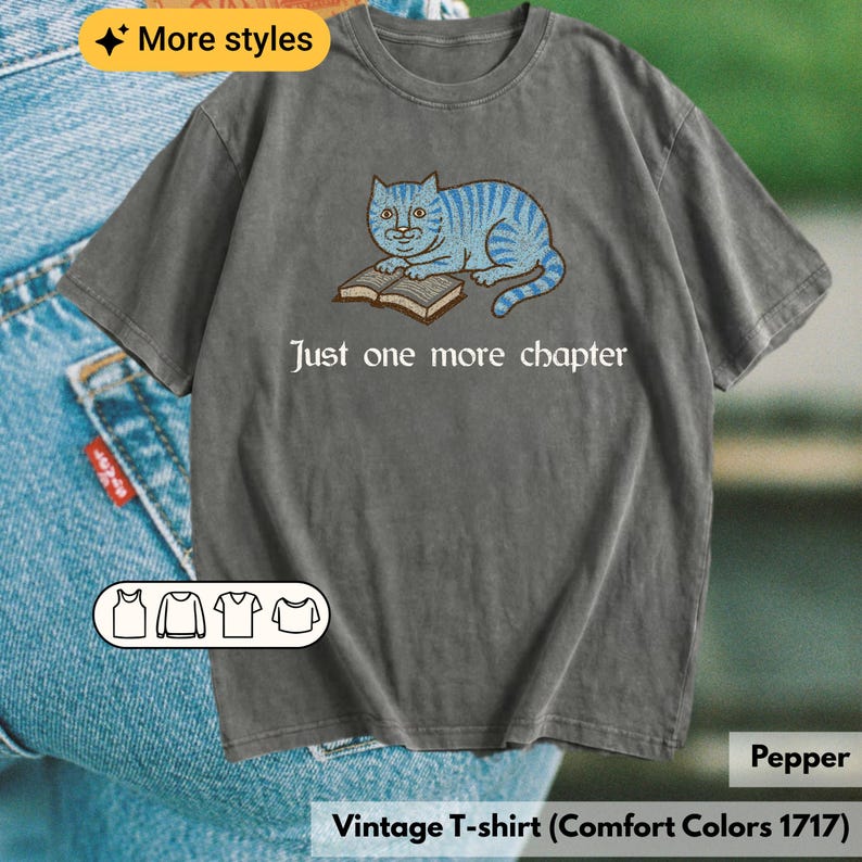 K&ouml;nnte beinhalten: Ein anthrazitfarbenes Kurzarm-T-Shirt mit einer Grafik einer blau gestreiften Katze, die mit einem offenen Buch daliegt. Unter der Katze steht der Text, Just one more chapter. Das T-Shirt hat einen Rundhalsausschnitt und eine Vintage-Stoffoptik.
