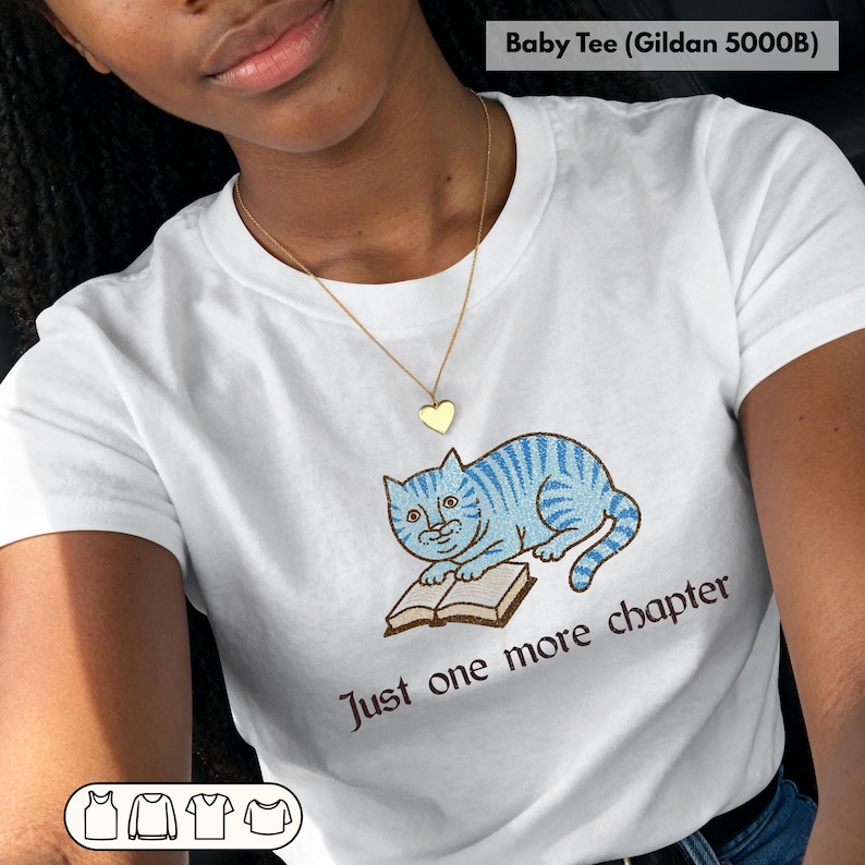 May include: White baby tee featuring a blue cat reading a book. The cat has blue stripes and is sitting on a brown book. The text "Just one more chapter" is below the cat in brown.