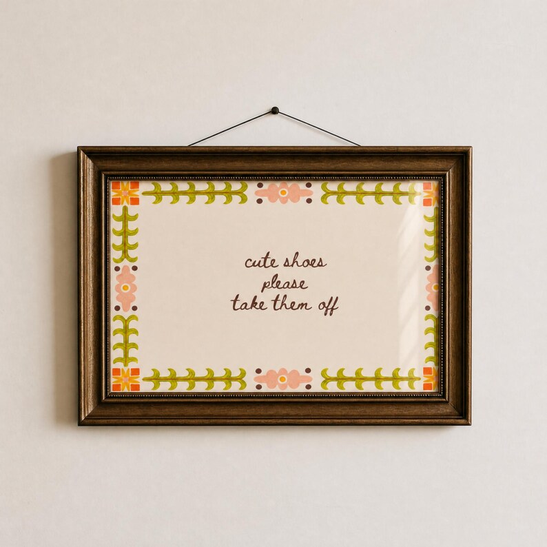 May include: A rectangular wall sign in a dark brown frame featuring a cream-coloured background with a decorative border of green leaves and pink flowers. The text in the centre reads, "cute shoes please take them off" in dark brown script.