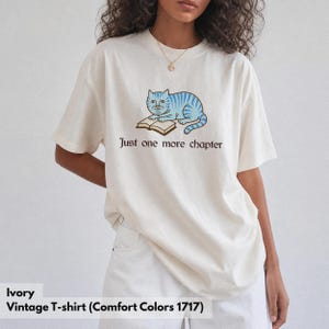 K&ouml;nnte beinhalten: Ein elfenbeinfarbenes Kurzarm-T-Shirt mit einer Grafik einer hellblau gestreiften Katze, die mit einem offenen Buch liegt. Unter der Katze steht der Text Just one more chapter in einer dunkelbraunen Serifenschrift. Das Shirt hat einen Rundhalsausschnitt.