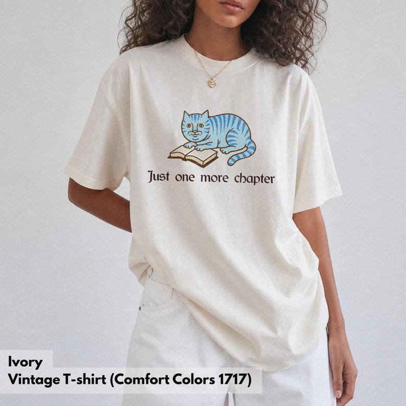 K&ouml;nnte beinhalten: Ein elfenbeinfarbenes Kurzarm-T-Shirt mit einer Grafik einer hellblau gestreiften Katze, die mit einem offenen Buch liegt. Unter der Katze steht der Text Just one more chapter in einer dunkelbraunen Serifenschrift. Das Shirt hat einen Rundhalsausschnitt.
