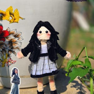 Könnte beinhalten: Eine handgefertigte Puppe mit schwarzen Haaren, einer schwarzen Jacke und einem weiß-grauen karierten Rock. Die Puppe hat einen hellen Hautton und trägt schwarze Stiefel. Der Hintergrund umfasst eine dekorative Vase mit Blumen- und Vogelmotiven.