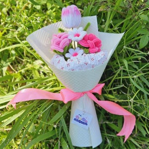 May include: A fabric flower bouquet featuring pink roses, white daisies, and a pink and white tulip. The flowers are wrapped in white paper and tied with a pink ribbon. A small tag is attached to the bouquet.