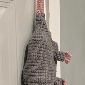 May include: A handmade crochet opossum doorstop in gray, white, and pink. The opossum is hanging upside down from a door handle. The doorstop has a gray body, white face, pink ears and paws, and a pink strap.