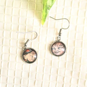 May include: A pair of silver-toned dangle earrings. Each earring features a round, glass-covered image of a vintage illustration. One depicts a boy in a red hat, the other a smiling face. The earrings hang from a simple hook.