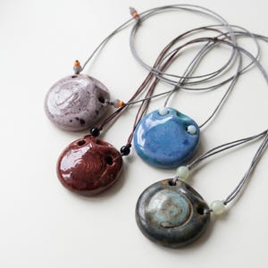 May include: Four ceramic pendants on thin gray cords, each featuring a unique glazed finish in shades of lavender, deep red, bright blue, and dark charcoal. Each pendant has a circular shape with small holes and decorative accent beads.