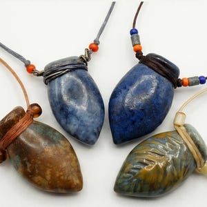 May include: Four teardrop-shaped ceramic pendants on cord necklaces. One is brown, one is mottled blue, one is deep blue, and one is green with a leaf pattern. Each pendant is secured with wrapped cord and small colorful accent beads.