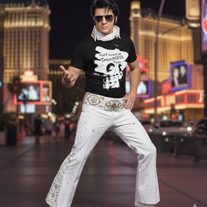 Puede incluir: Una persona vestida como Elvis Presley, con una camiseta negra con el texto "Make America Trumpless Again", pantalones blancos con tachuelas decorativas, un cinturón blanco y gafas de sol. El fondo muestra una ciudad de noche.