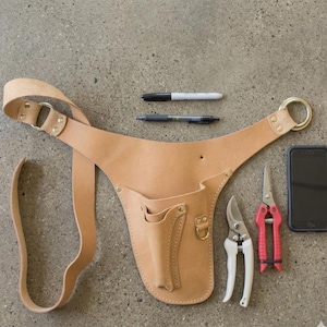 May include: A tan leather gardening tool belt with pockets, a gold ring, and a strap. Two pens, a black marker, a pair of white and red handled pruning shears, and a smartphone are also visible.