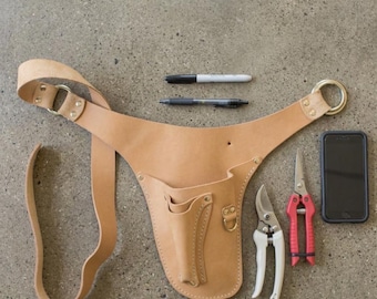May include: A tan leather gardening tool belt with two pockets, a pen, a marker, two pairs of pruning shears, and a smartphone. The shears have white and red handles. The belt has a gold-colored ring.