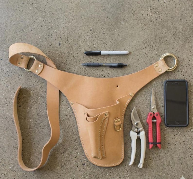 May include: A tan leather gardening tool belt with pockets, a gold ring, and a strap. Two pens, a black marker, a pair of white and red handled pruning shears, and a smartphone are also visible.