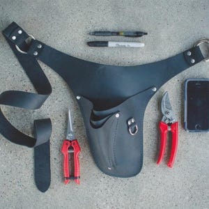 May include: Black leather gardening tool belt with two red-handled pruning shears, a smartphone, and writing utensils. The belt has a loop for hanging and multiple pockets for tools.