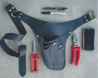 May include: Black leather gardening tool belt with two red-handled pruning shears, a pair of scissors, a smartphone, and writing utensils. The belt has a loop for hanging and a long strap.