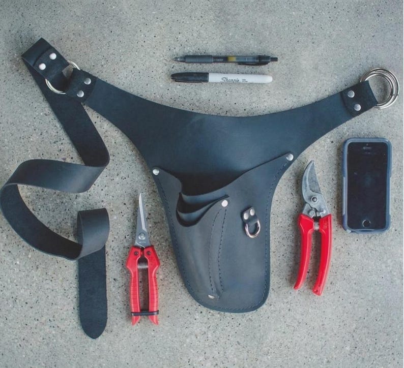 May include: Black leather gardening tool belt with two red-handled pruning shears, a smartphone, and writing utensils. The belt has a loop for hanging and multiple pockets for tools.