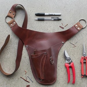 May include: A brown leather gardening tool belt with a shoulder strap, two metal rings, and multiple pockets. Two pairs of red handled pruning shears and two pens are also visible.